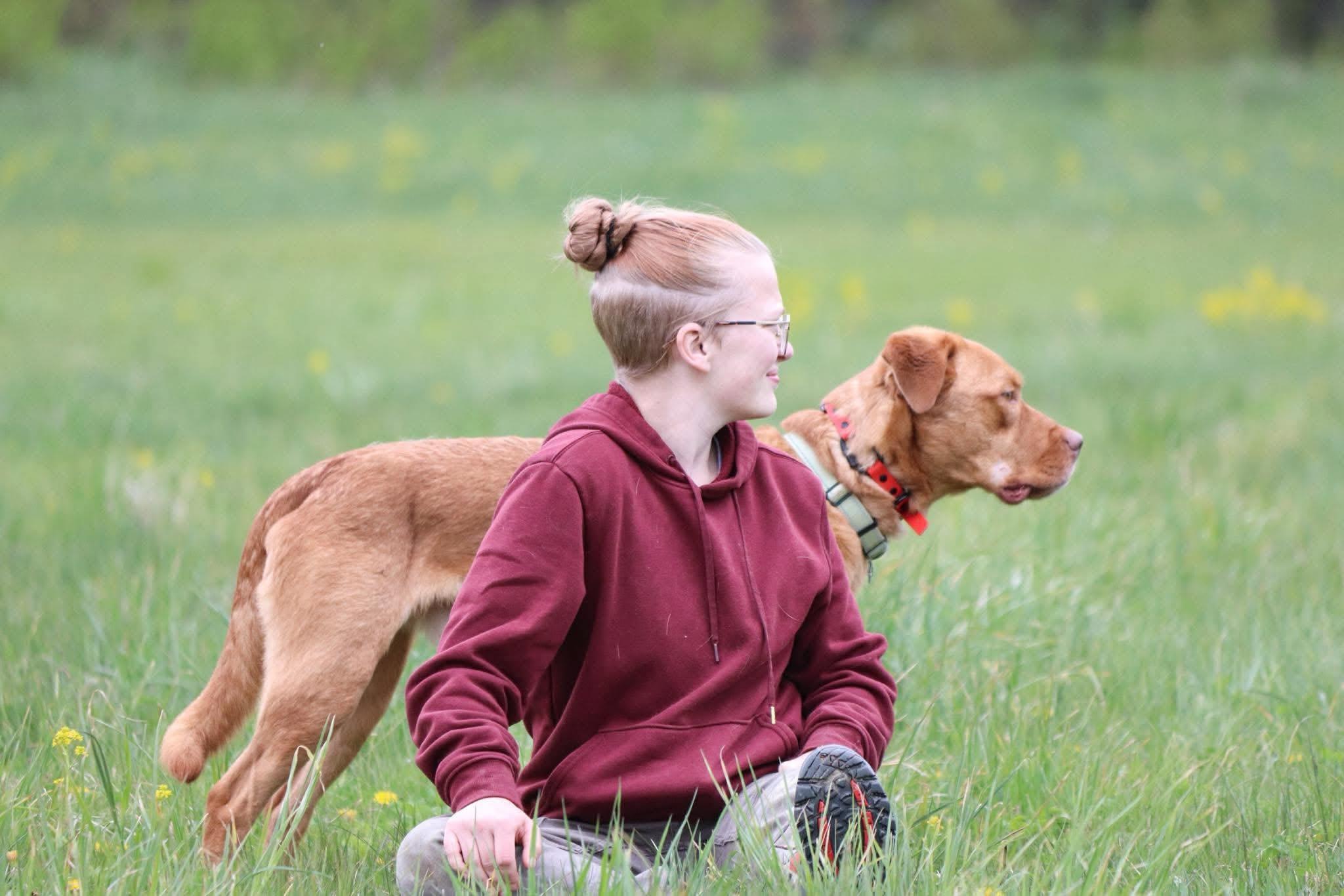 Savannah kneeling in a sunlit grassy field beside a relaxed tan dog
